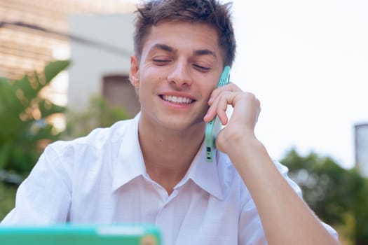 Happy young man talking on a smartphone during a phone call outdoors. He is smiling and looking away, suggesting a positive conversation in a modern and relaxed urban environment with sunlight.