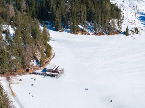 Frozen Braies Lake in the Dolomites, Italy, surrounded by evergreen trees. A wooden boathouse with a walkway sits on the ice, with people nearby.