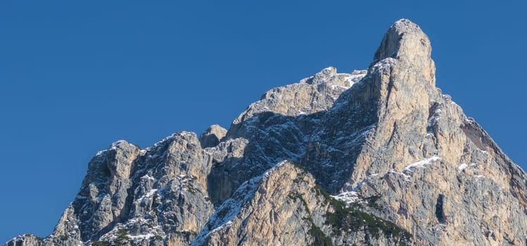 A rugged mountain peak in the Dolomites, northern Italy, with rocky formations partially covered in snow, set against a clear blue sky.