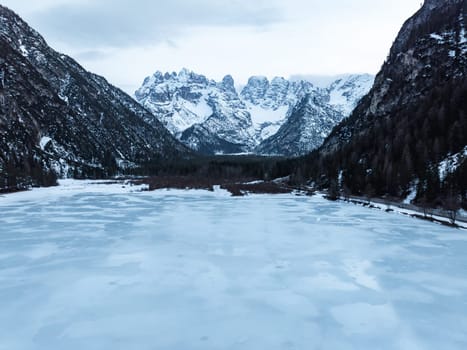 A frozen lake with an icy surface lies amidst the towering snow covered peaks of the Alps, with dense forests along the lower mountain slopes.