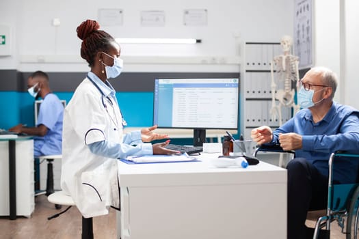 Black doctor uses computer to review caucasian pensioner patient medical records. Female physician with stethoscope listens carefully as retired male wheelchair user describes his health concerns.