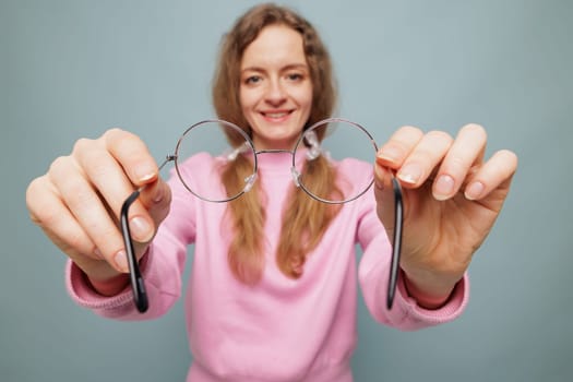 A smiling woman in a pink sweater holds a pair of round glasses towards the camera, set against a light blue background.