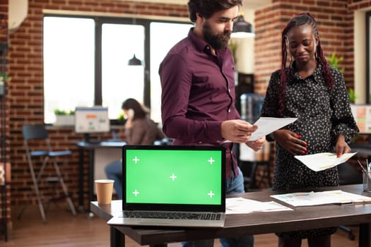 Laptop with greenscreen placed on table as pregnant woman and caucasian man review business paperwork. African american expectant mother and male colleague working as device displays mockup template.