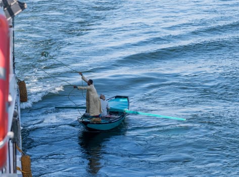 Esna, Egypt - 3 November 2024: Street vendor in a small rowboat selling textiles throwing rope to attach to a passing cruise ship.