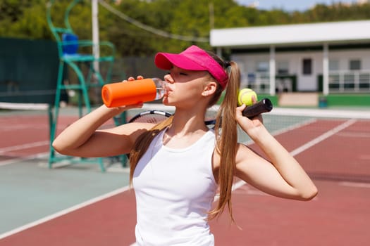Young female tennis player drinking water, holding racket and ball, wearing sportswear and visor, rehydrating after practice on an outdoor court, tennis game