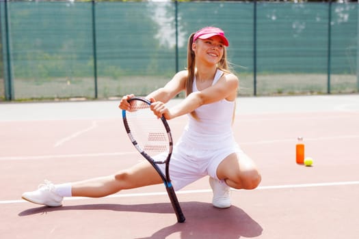 Smiling female tennis player stretching on court, using racket for balance, wearing sportswear and visor, preparing for training in sunny weather, tennis game