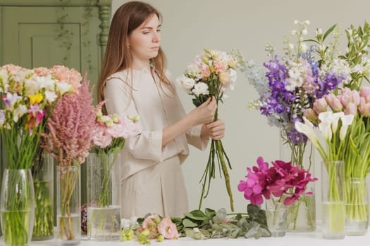 Female florist creating a delicate bouquet with fresh flowers in a floral workshop, surrounded by colorful blooms in glass vases. flower shop