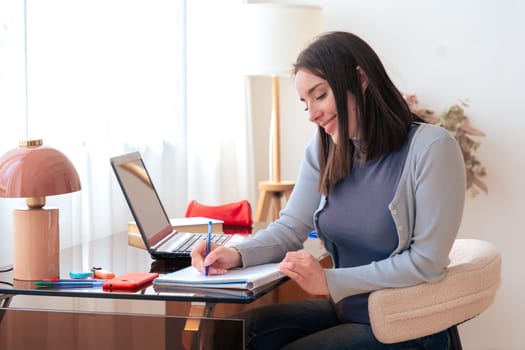 Caucasian smiling student studying at home, writing in notebook while using laptop. Modern workspace with stylish decor, lamp, and stationery. Education, learning, productivity, and concentration.
