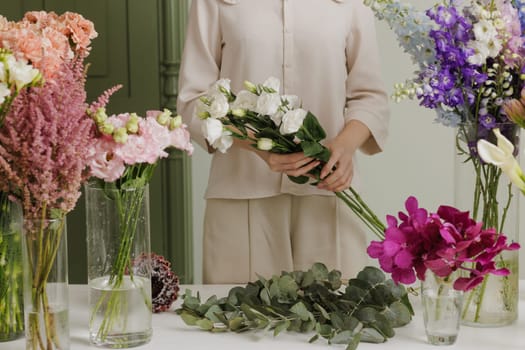 Female florist creating a delicate bouquet with fresh flowers in a floral workshop, surrounded by colorful blooms in glass vases. flower shop