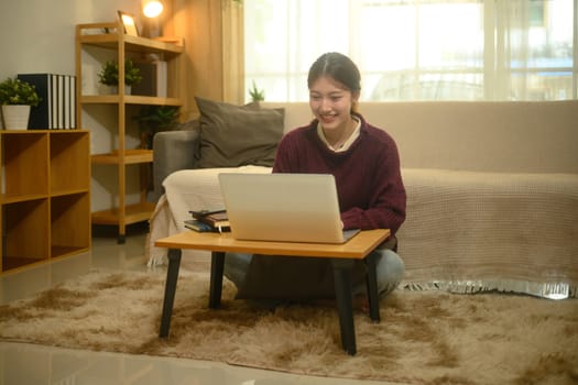 Smiling asian female freelancer working with her laptop, sitting on carpet in living room.