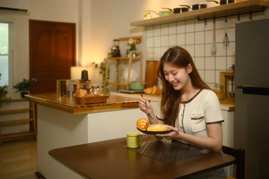 Happy woman sits at a kitchen table, enjoying a delicious breakfast of eggs and croissant.