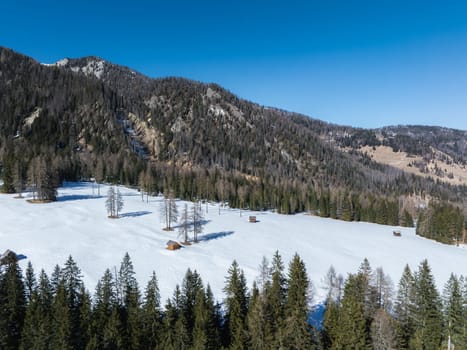 Snow covered field with scattered wooden huts, surrounded by dense evergreen forests and rugged mountain slopes under a clear blue sky near Braies Lake.