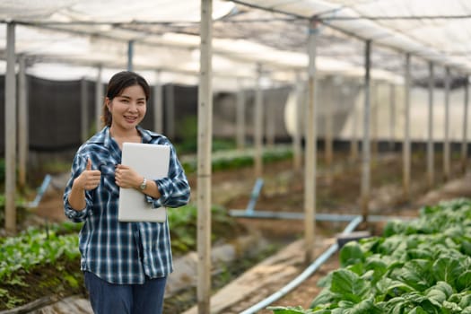 Confident female farmer stands in a greenhouse holding laptop and giving a thumbs up.