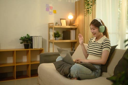 Cheerful asian woman wearing headphones, smiling and waving during virtual call in a cozy home.