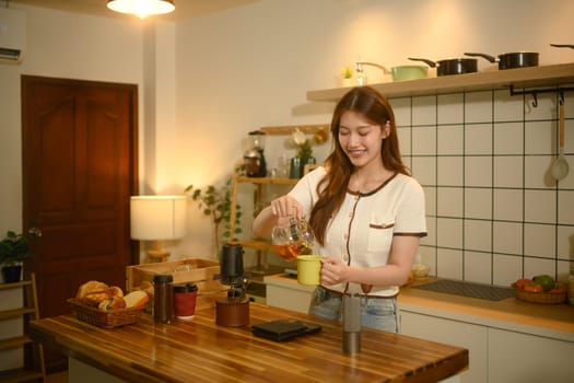 Happy asian woman pouring freshly brewed coffee into a mug in her warm kitchen.