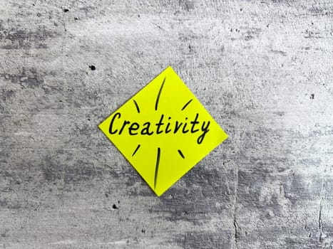 Vibrant yellow sticky note shaped as a diamond displaying the word Creativity and decorative elements on a textured grey background, representing the essence of innovative thinking and inspiration