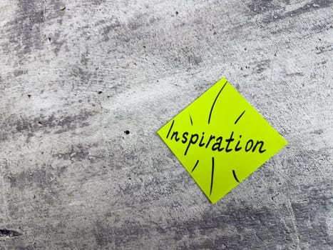 Vibrant yellow sticky note in a diamond shape displaying the word Inspiration with decorative elements on textured grey surface, representing the essence of motivating ideas and creative potential