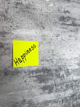 Bright yellow sticky note inscribed with the word Happiness, displayed on a textured grey background, representing the essence of joy and the pursuit of a fulfilling, positive life