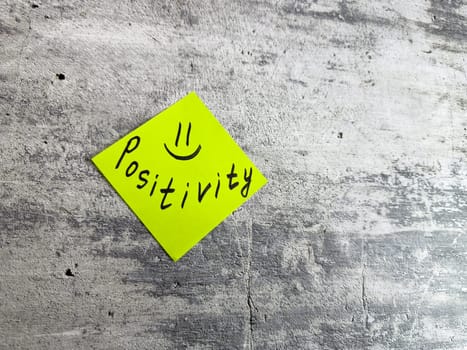 Bright yellow sticky note shaped as diamond featuring the word Positivity and smiley face on textured grey background, symbolizing cheerful mindset and the power of positive thinking in daily life