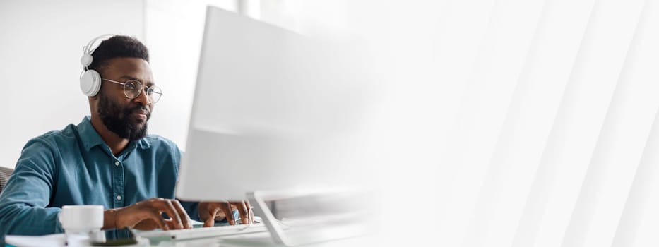 A young African American man works diligently at his desk in a modern office. He wears headphones and is engaged in tasks, showcasing a successful lifestyle as a manager and entrepreneur, copy space