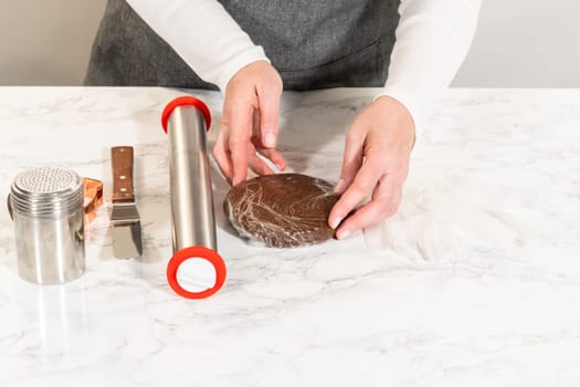 Front-facing view of hands unwrapping chocolate gingerbread dough on a marble surface, with baking tools and a mitten-shaped cutter nearby.