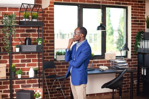 African american male manager standing in brick wall office and looking at different plants arranged on cabinets. Black man with hand on his chin, contemplating on business ideas in empty workspace.