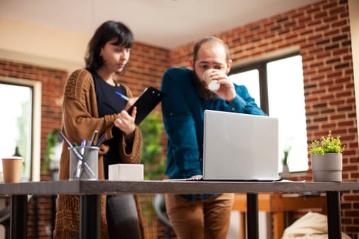 Bearded man and young woman collaborate on laptop, discussing business project goals. Female employee with clipboard and male coworker drinking coffee are looking at device screen in startup company.