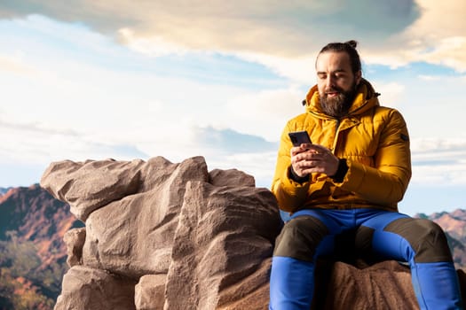 Nature lover relaxing on mountain top getting online with his phone at a scenic viewpoint, looking out over the landscape. Hiker staying always online and connected, panoramic scenery.