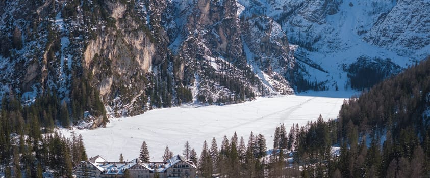 Aerial view of Braies Lake in winter, surrounded by rugged cliffs and evergreen forests. A lodge is visible near the frozen lake with faint tracks on the ice.