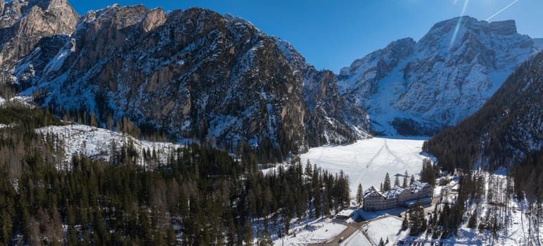 Aerial view of Braies Lake in winter, surrounded by snow covered peaks, evergreen trees, and a lodge near the frozen shoreline under a bright blue sky.