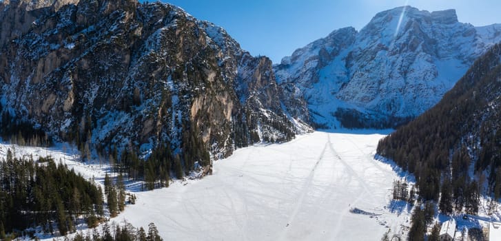 Aerial view of Braies Lake in winter, frozen and snow covered, surrounded by rugged mountains, dense forests, and a clear blue sky.