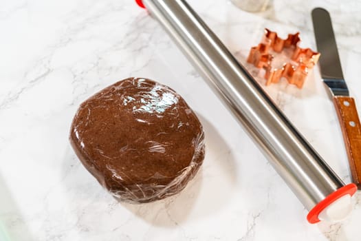 A ball of Chocolate Ginger Sugar Cookie dough wrapped in plastic, placed on a marble surface with baking tools in the background. Close-up perspective.