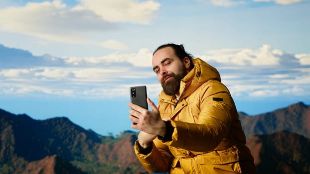 Determined adventurer enjoys the panoramic mountain landscape, taking selfies with the amazing scenic background on the hill top where nature and towering peaks create a breathtaking view. Camera B.