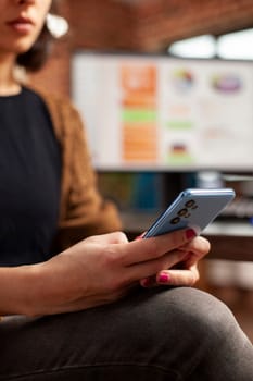 Young businesswoman sitting her desk, holding cellphone to create her schedule. Closeup of female employee using mobile device, checking messages and social media in startup office.