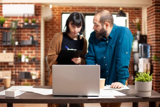 Caucasian businesswoman and man standing at table, discussing marketing strategies with laptop open. Female employee with clipboard and coworker are focused and committed to achieving business goals.