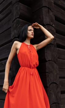 A slender woman in a vibrant red dress shields her eyes, gazing towards the horizon against a rustic, dark wooden wall.