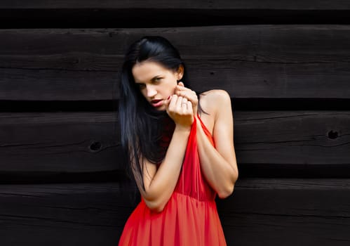 A slender woman in a vibrant red dress shields her eyes, gazing towards the horizon against a rustic, dark wooden wall.