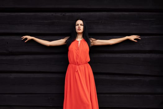 A slender woman in a vibrant red dress shields her eyes, gazing towards the horizon against a rustic, dark wooden wall.