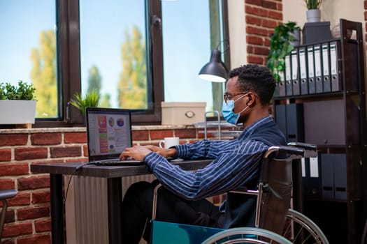 Male wheelchair user types on his personal computer, updating and reviewing project development plans in startup office. Paralyzed entrepreneur using laptop and analyzing business analytics at desk.