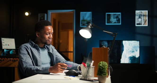 Casual guy purchasing products on electronic app with his credit card, ordering apparel on internet during promotional season. Black man feels satisfied as he enters payment information. Camera A.