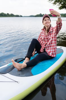 A woman in a red plaid shirt and leggings sitting on a paddleboard, smiling while taking a selfie on a calm lake.