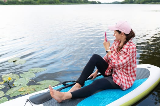Woman relaxing on a SUP board on a calm lake, wearing a cap and plaid shirt, using her smartphone.