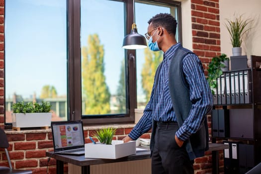 Business advisor wearing face mask stands at desk, arranging a box of work materials, including a plant pot. Black entrepreneur is reviewing important business project details and preparing to leave.