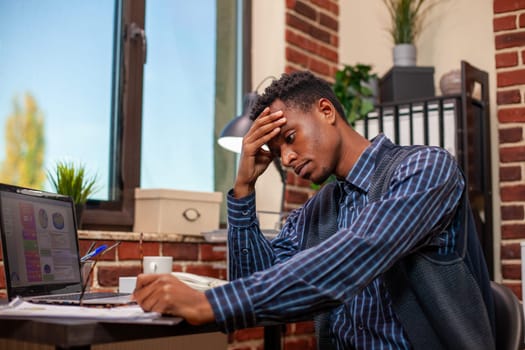 African american businessman leans on table, frustrated while working on marketing strategy presentation on laptop. Black male employee seated at desk with forehead in his hands, overwhelmed by work.