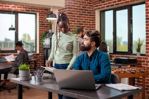 Black businesswoman with a clipboard discusses marketing strategy ideas with male colleague in brick wall workspace. Caucasian bearded man listens intently to female coworker at his desk in office.