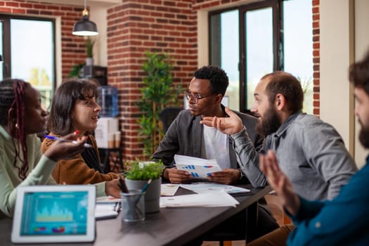African american male manager conducting group discussion with his employees in startup office. Colleagues sitting together in brick wall workspace, having a business meeting with documents on table.