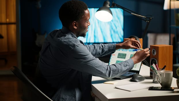 Black man organizing his schedule with a planner and putting a reminder, creating an adhesive note post it and browsing the web. Modern productive guy writes a memo for research. Camera B.