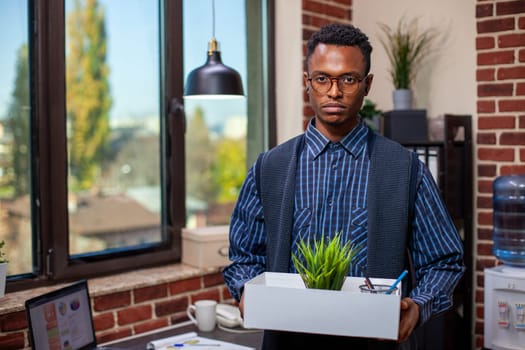 Portrait of black male employee holds a box containing green plant and stationery, standing in brick wall workspace. Businessman with a white carton of office materials, looking at camera.