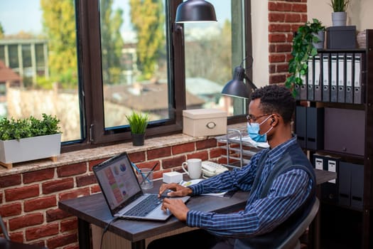 Black startup employee with face mask, wearing wireless earbuds and reviewing project insights on personal computer. Male analyst listening to music and working on laptop in brick wall office.