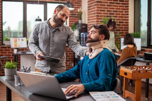 Injured analyst seated at desk with his laptop, receiving friendly support from coworker in office. Male manager stands with clipboard and speaks to his employee with a neck brace in modern workspace.